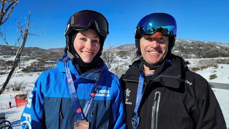A man and a woman in snow gear with medals on a mountain top.