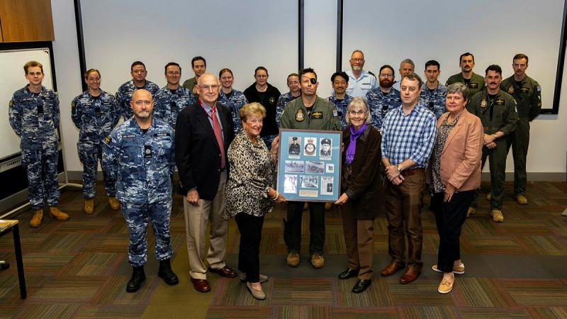 A group of military personnel and civilians stand in a group with a large blue plaque.