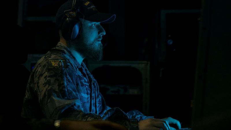 A Navy member sits in a darkened room, with a blue light from a screen partially illuminating him.