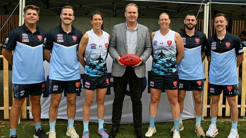 Four men in sports uniform, two women in sports uniform and a man in a suit and holding an AFL ball stand in a line.
