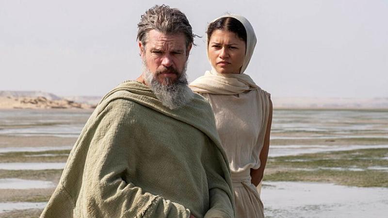 A haggard old man and a young woman stand in beige, flowy clothes in a flat, puddled plain.