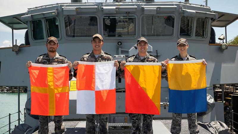 Four Navy personnel from HMAS Diamantina hold up signalling flags on the deck of the ship.