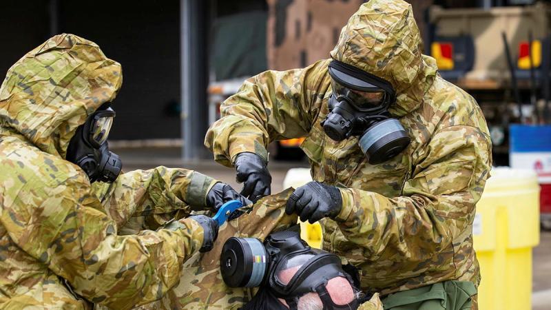 Two people in hazmat suits reach under the hood of a third person in a hazmat suit who is lying down.