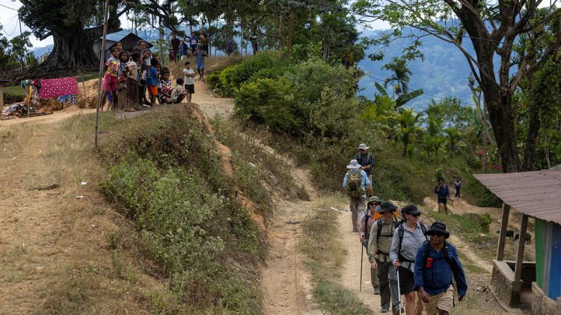 People wearing backpacks walk up a dirt track past a small hut as children watch from the ground above.