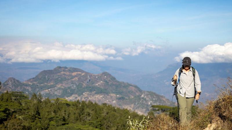 A woman wearing a baseball cap stands on a peak overlooking a valley.