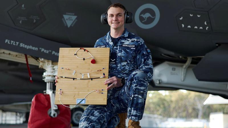A military man wearing headphones holds up a board affixed with wiring.
