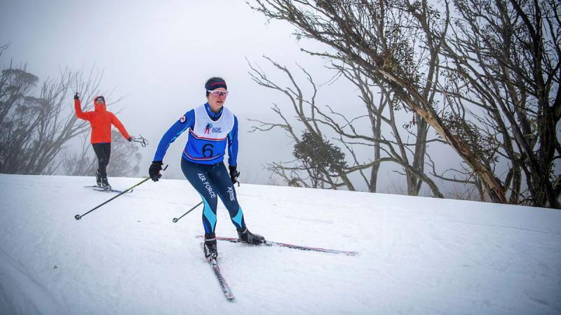 An Air Force woman on skis paces another skier in an orange coat on the slopes.