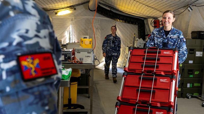 A woman in an Air Force uniform pushes a trolley stacked with red boxes as two other women stand and watch.
