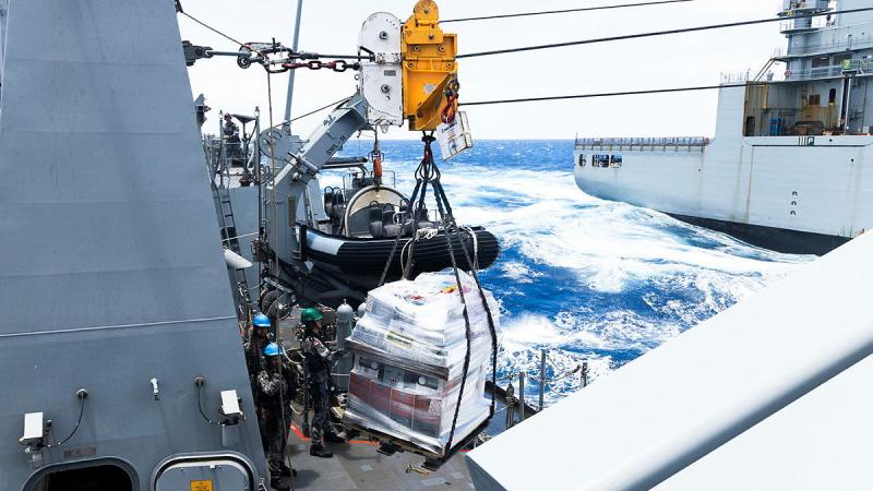 A large parcel of goods hangs from cables strung between two ships at sea. 