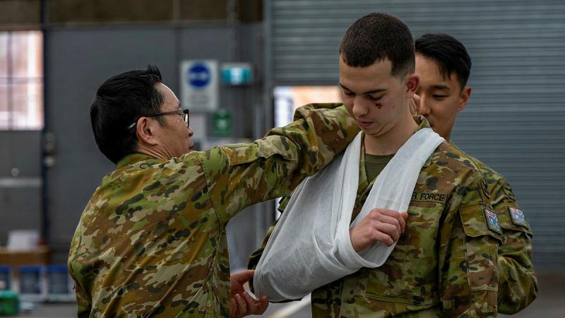 An Australian Air Force Personnel member ties an arm sling on a fellow officer.