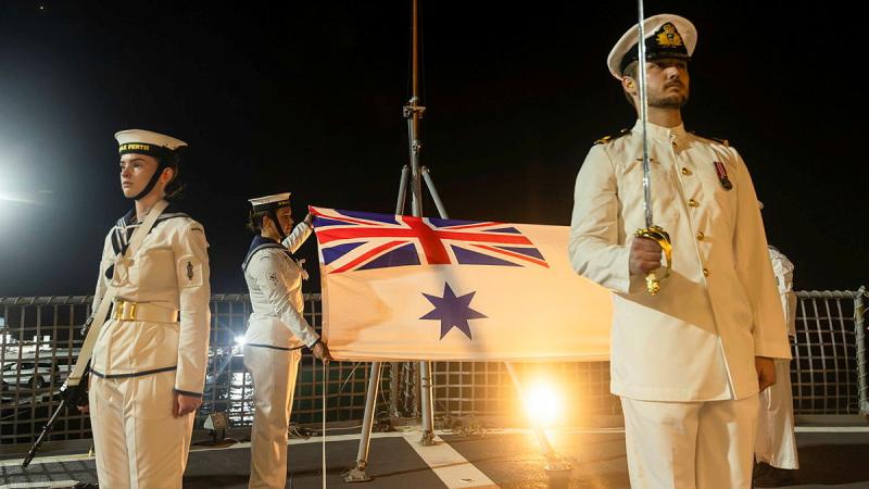 A night sky is lit, showing Navy personnel in white uniforms standing with a flag, sword and gun.