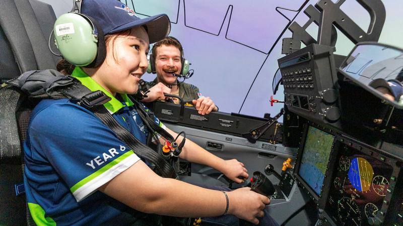 A child flies an aircraft simulator, guided by a military man.