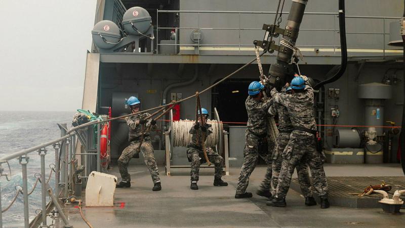 Navy personnel in blue hard hats reach for lines and other equipment on the deck of a ship.