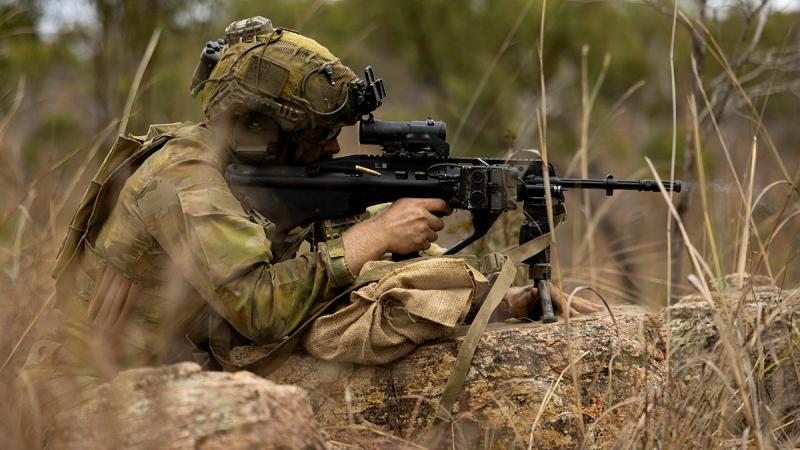 A soldier aims a gun while crouched behind a log.