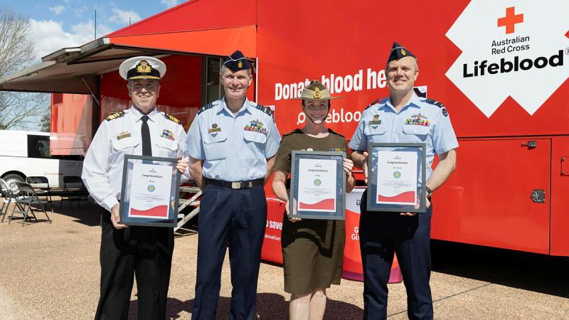 Four military personnel stand outside a blood donation van, three with certificates.