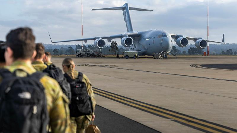 Military personnel with backpacks walk across the tarmac to a stationary aircraft.