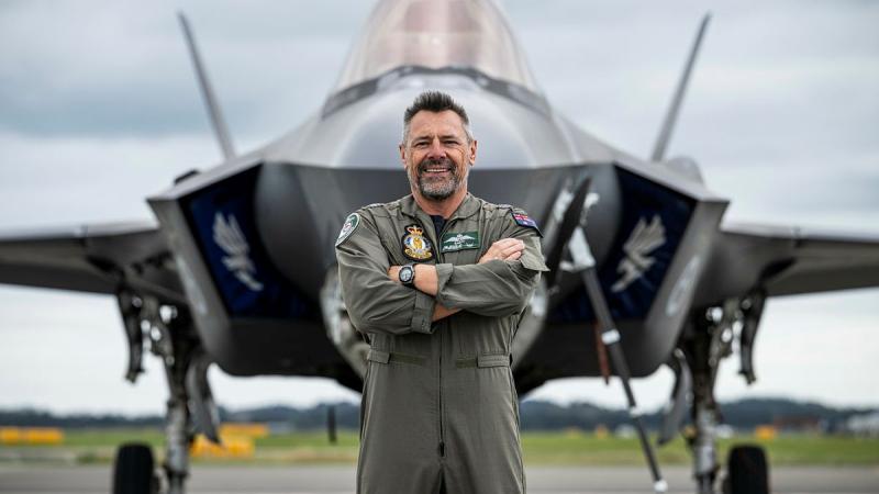 A military man in a flight suit stands before an aircraft on the tarmac.