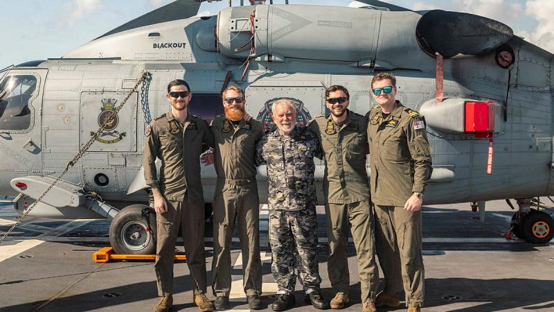 Five people dressed in a mix of flight uniforms and Navy cams stand in front of a helicopter.