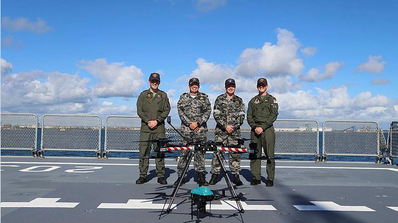 Four people in uniform stand next to a drone on a stand.