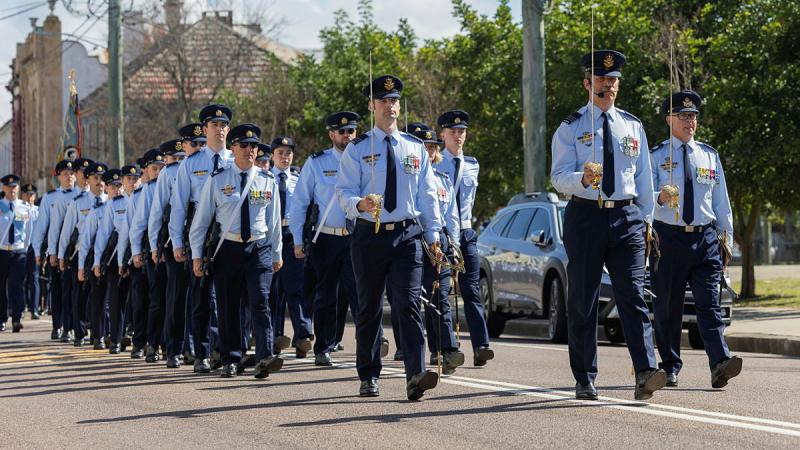 A large group of Air Force personnel with swords held in the air, march together through a city street.