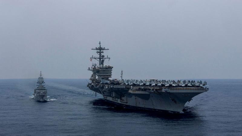 A ship sails close to a much larger vessel which carries numerous aircraft.