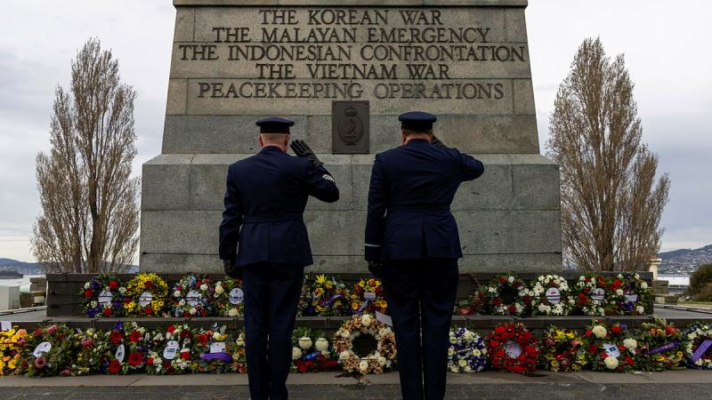 Two military men salute a collection of wreaths at the base of a memorial for several wars/operations.