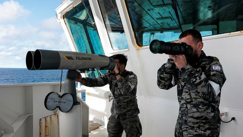 Two people in Navy uniforms look through very long and large lenses from the deck of a boat.
