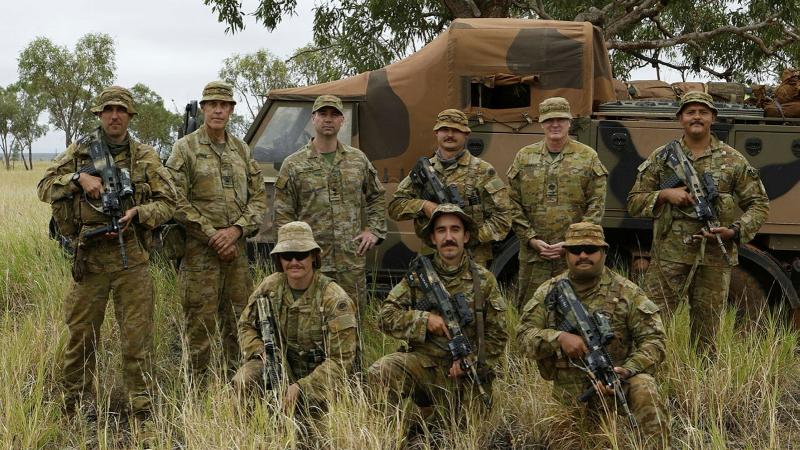 A group of soldiers stand with weapons in front of an Army vehicle in a bush setting.