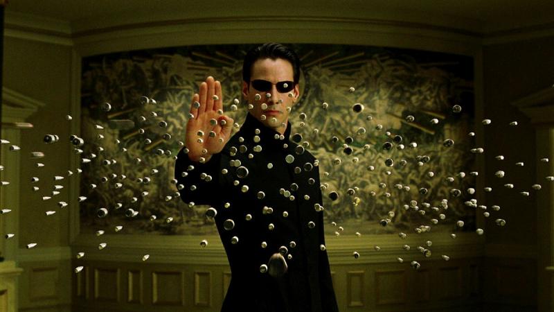 A man wearing sunglasses holds his hand up in front of him as bullets head towards him.