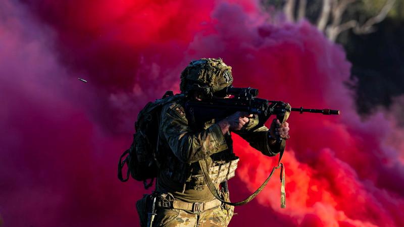 A person in Army uniform shoots a rifle, with the shell flying out behind them, against a backdrop of pink smoke.
