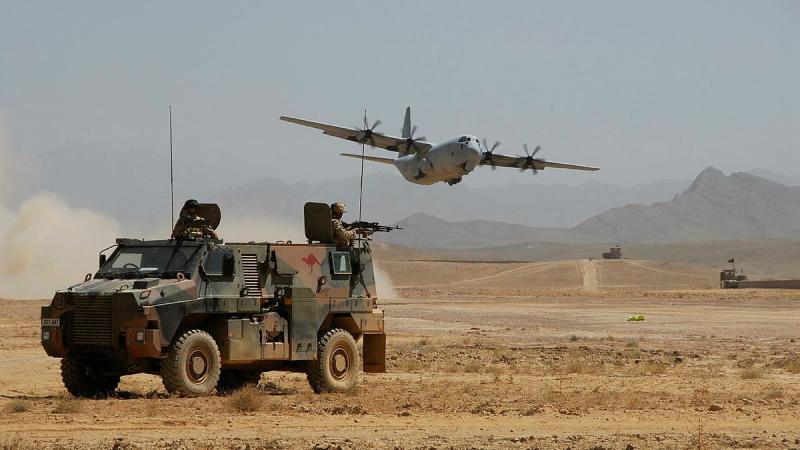 An Army vehicle is parked in the foreground on a dirt plain while an aircraft flies overhead