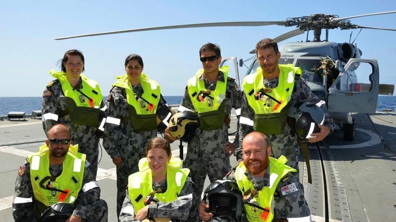 Seven military personnel wear life vests while on a ship at sea, with a helicopter behind them.
