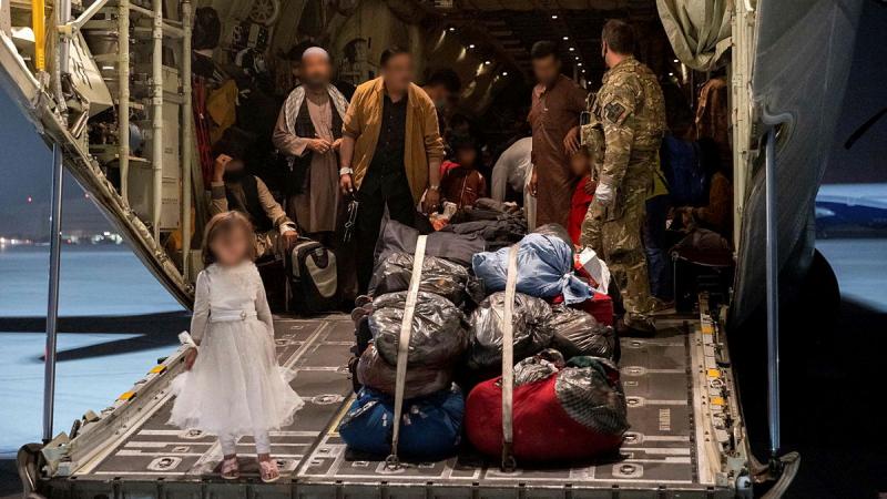A child stands on the ramp of an aircraft beside a bundle of filled bags, with a group of people further back in the aircraft