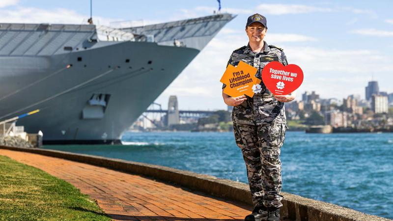 A Navy member in uniform stands with a red heart-shaped sign and a yellow cross-shaped one near a ship and water, with city buildings in the background.