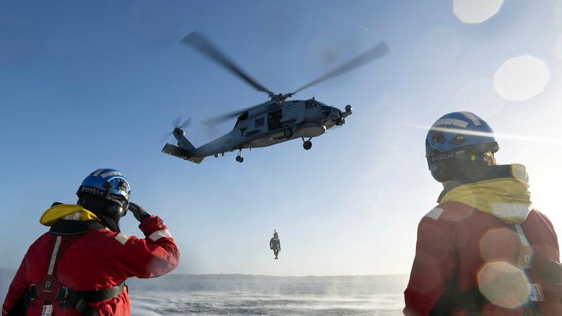 A helicopter hovers over water with a person suspended from a winch underneath while two people in helmets and orange jackets watch.