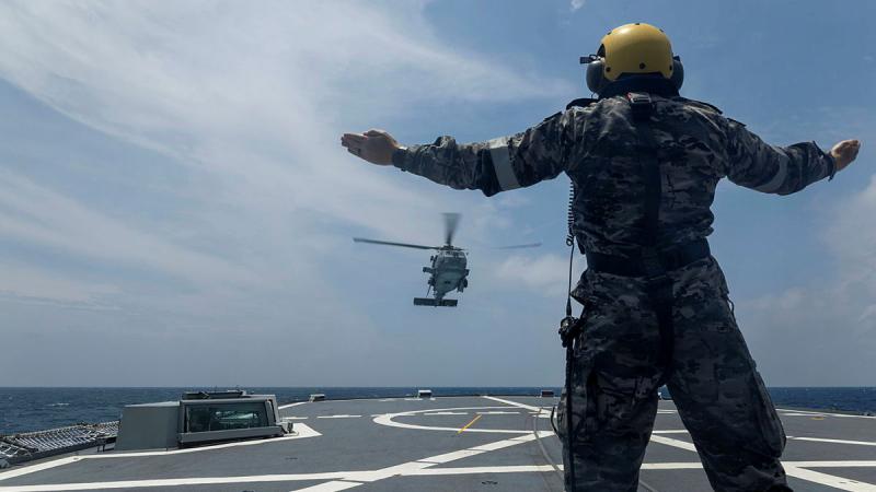 A military man wearing a helmet signals with his arms to an incoming helicopter, while onboard a ship at sea.