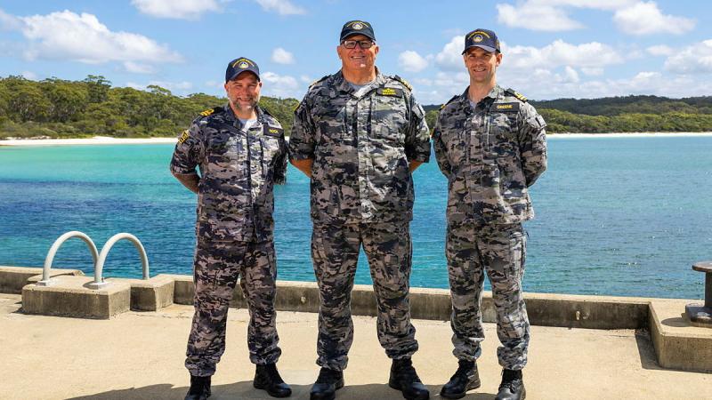 Three men in Navy uniform stand side by side with their backs to a bay, with a tree-lined shore in the background.