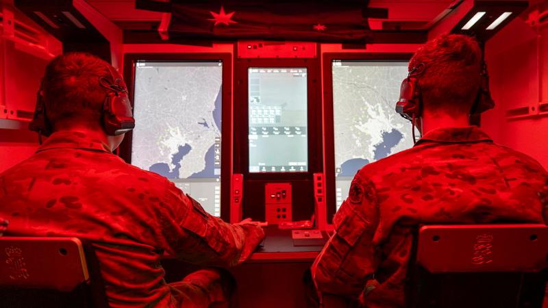 Two people in uniform sit looking at screens in a small room with red lighting.