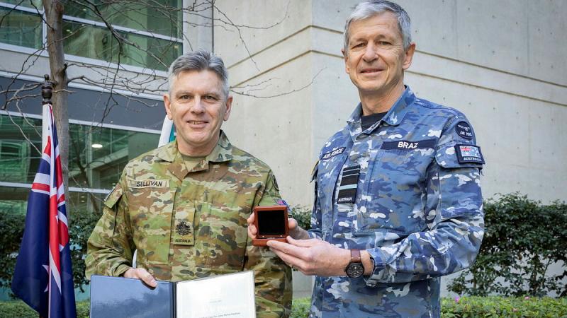 Two men in uniform stand side by side outdoors next to an Australian flag, one holding a certificate, the other a medallion box.