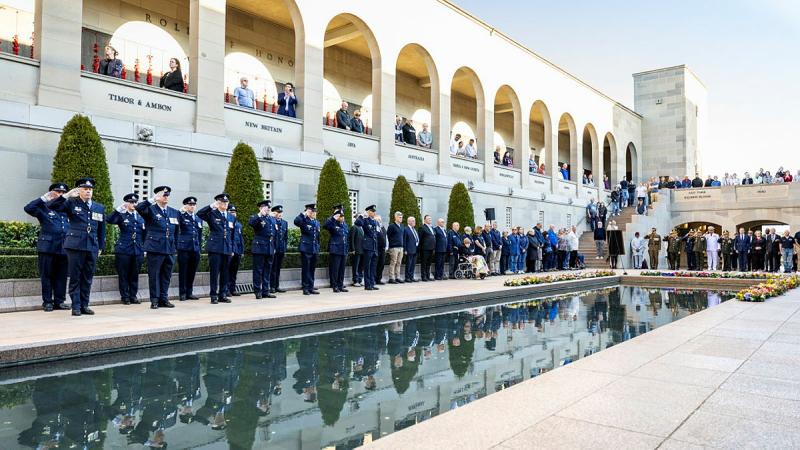People in uniform salute beside a long reflective pool as people watch from balconies above.