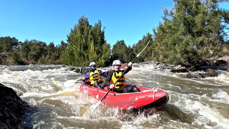 Two people in life vests and helmets paddle through water while on board a red raft.