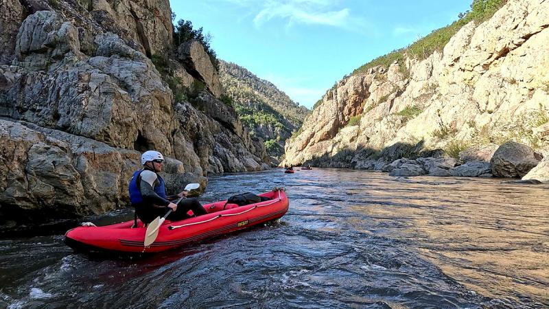 A person wearing a helmet and in a kayak floats on a river flanked by high rocky terrain.