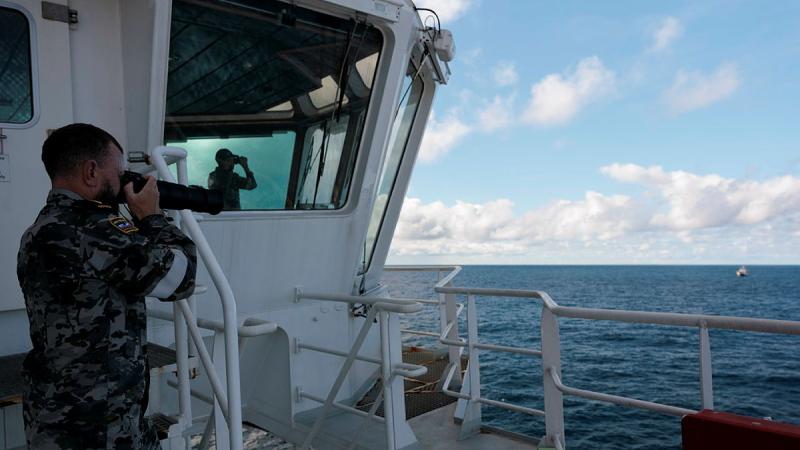 A man in a Navy uniform holds a camera up to his eyes as he looks towards a boat at sea.