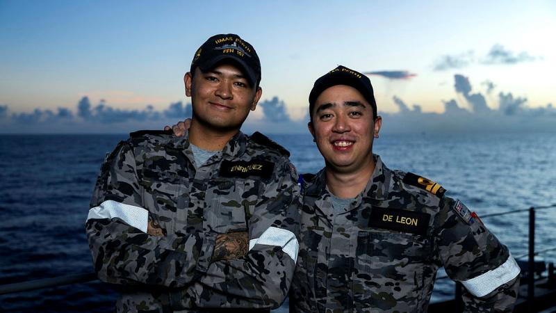 Two men in Navy uniform stand side by side on the deck of a ship with the sea behind them.