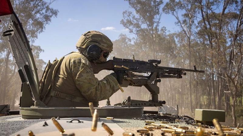 A person in uniform fires a mounted gun out of the hatch of a vehicle, with bullet shells scattered across the vehicle roof.