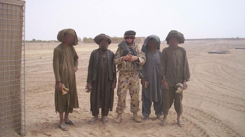 A military man stands with four civilians, whose faces have been blurred out, in the desert.