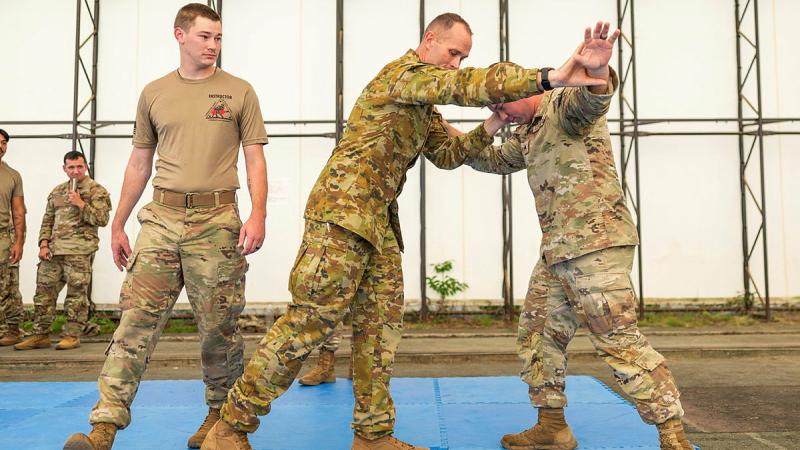 Two men perform a martial arts move on a gym mat while another observes behind them.