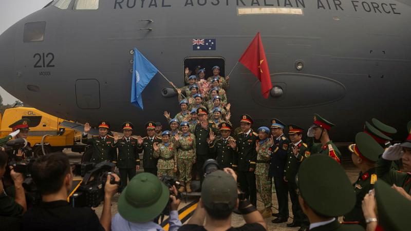 A group of military personnel stand in front of an aircraft with flags, while waving to a crowd.
