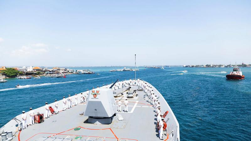 Sailors line the deck of a ship as it sails into a port.