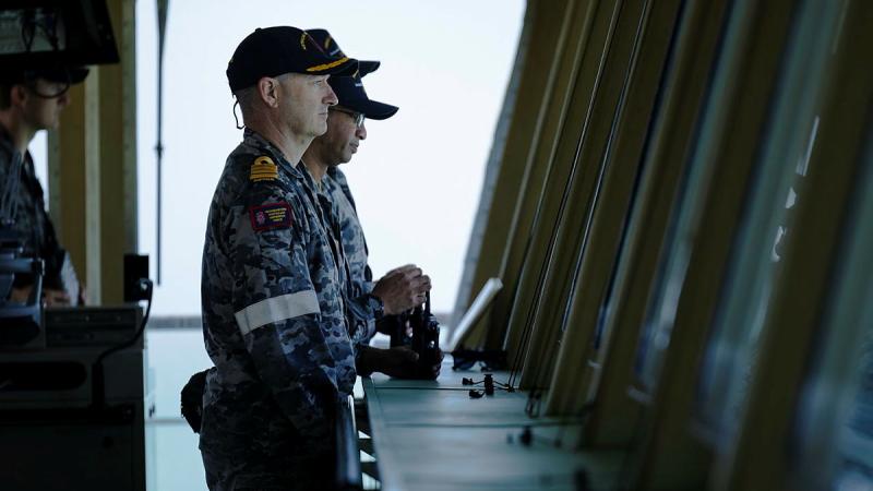 Navy members stand side by side on a ship staring out to the ocean.